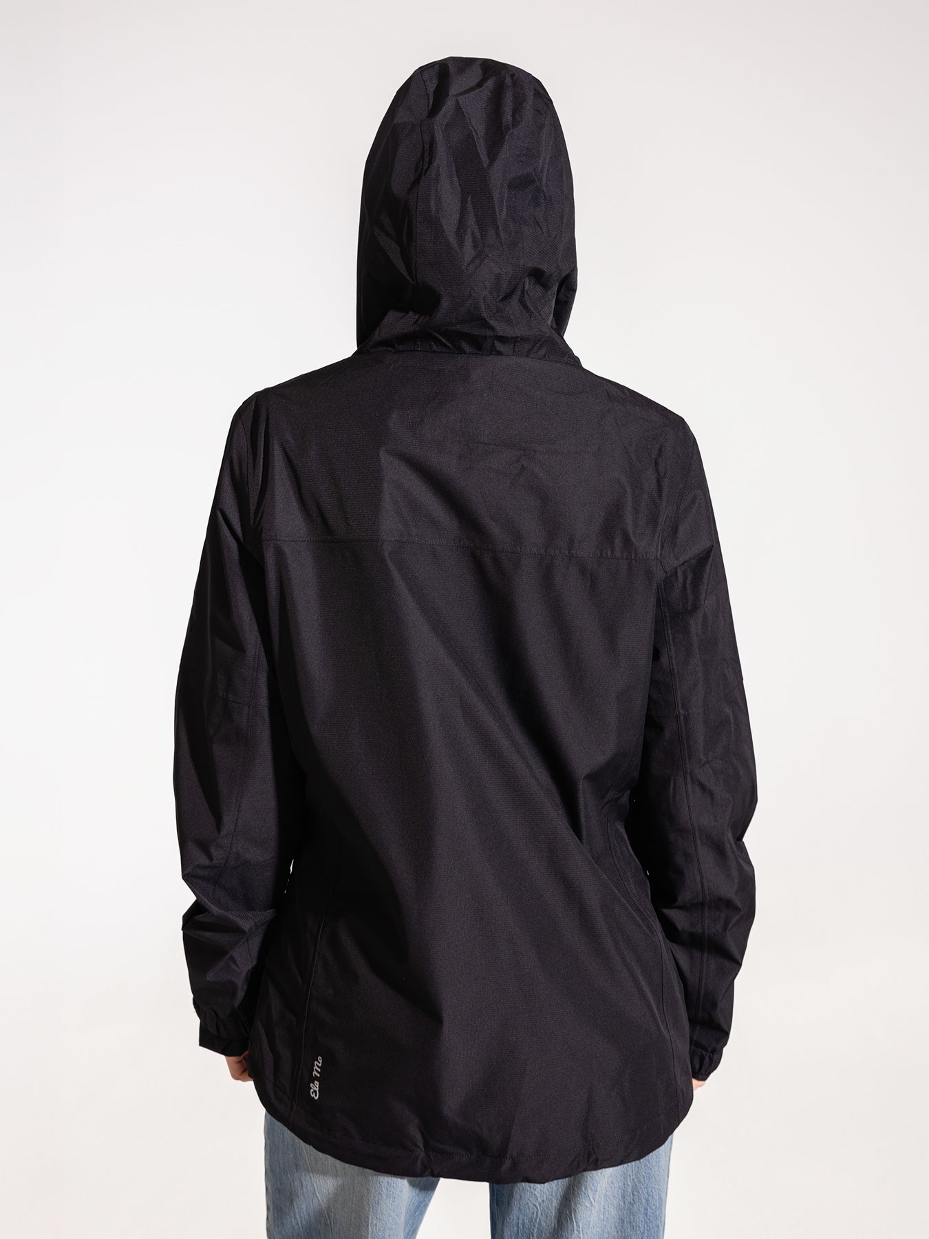Ela Mo™ Regenjacke | Black. Eine Frau trägt die schwarze Regenjacke, Kapuze auf dem Kopf, Rückenansicht.