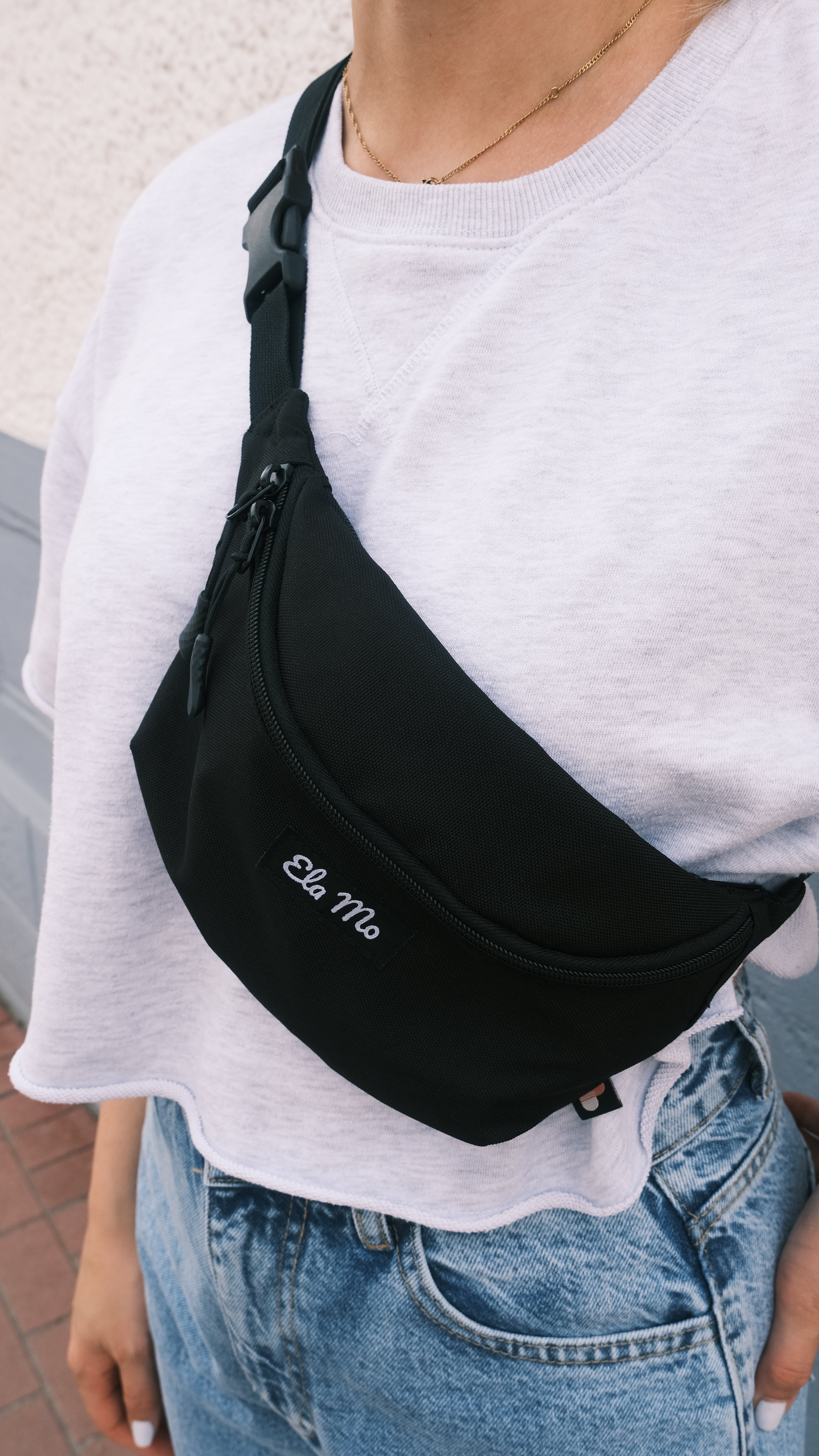 Ela Mo™ Hipbag | Black. Eine Frau mit Hipbag auf weißem Shirt, alltäglicher Lifestyle.