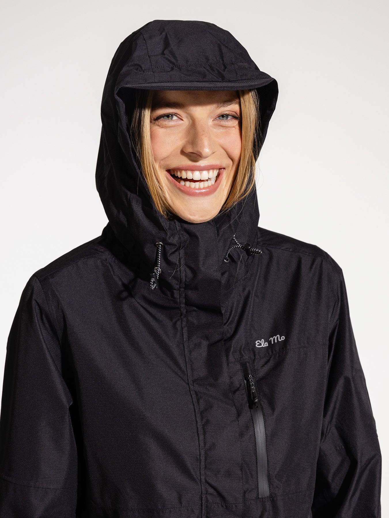 Ela Mo™ Regenjacke | Black. Eine blonde Frau mit schwarzer Regenjacke und Kapuze auf dem Kopf.