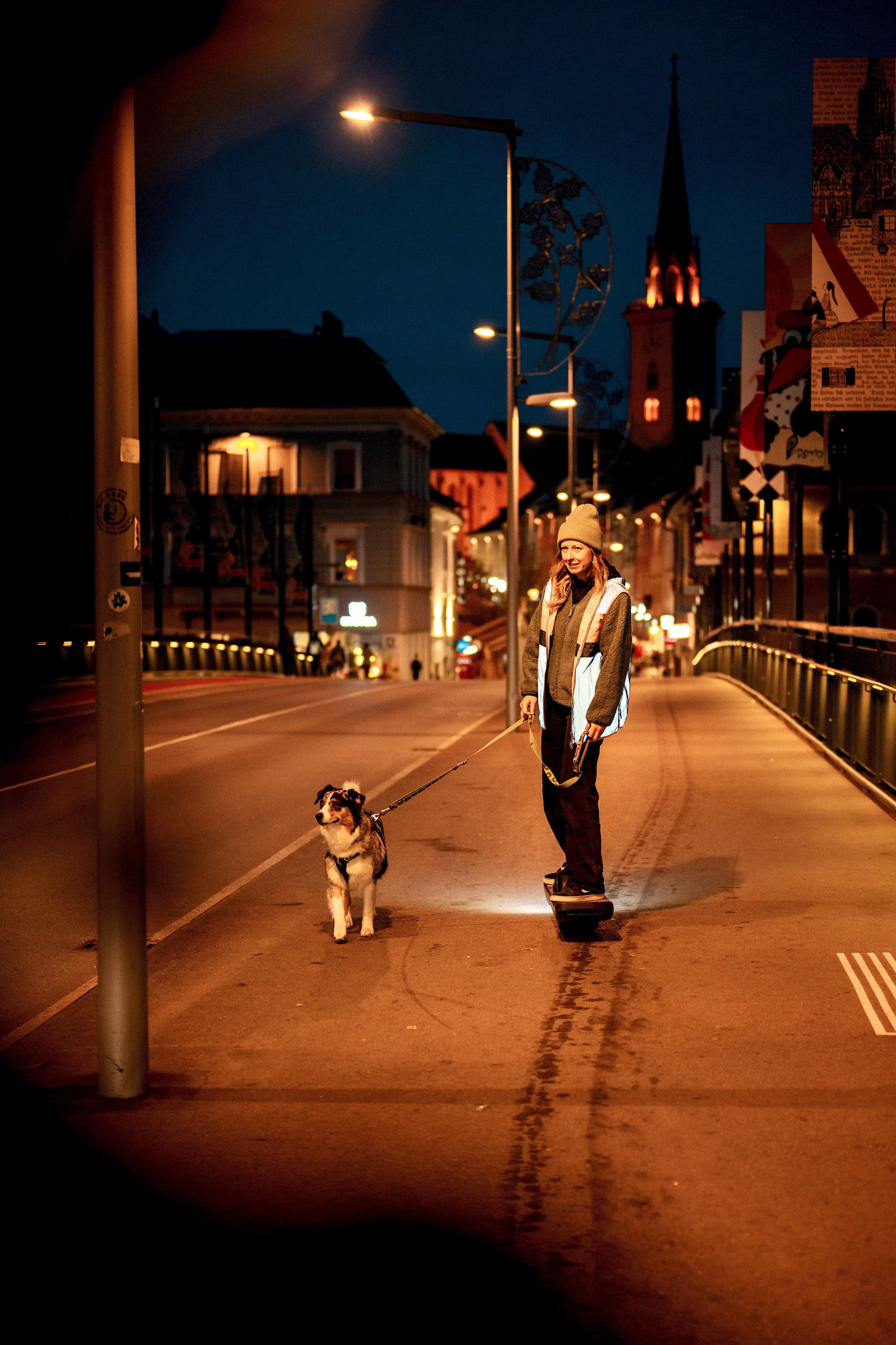 Ela Mo™ Fahrradweste.Eine Frau in reflektierender Fahrradweste in der Nacht mit Hund und Skateboard.