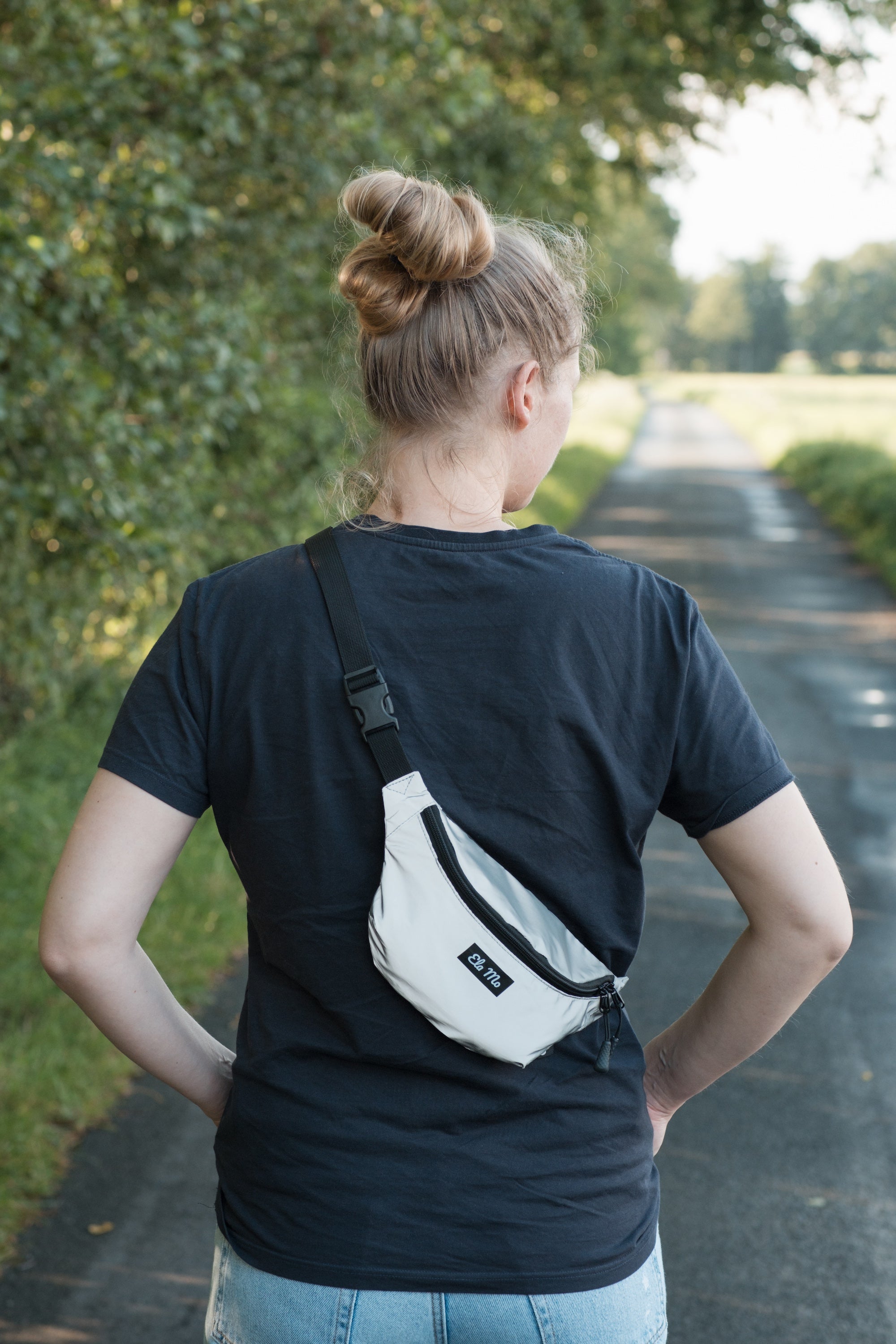 Ela Mo™ Hipbag  | Reflective. Eine Frau mit Hipbag an einem sonnigen Tag auf der Wiese, Ansicht von hinten.