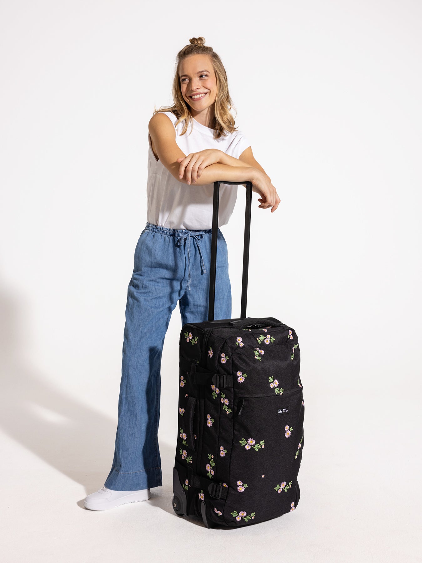 Ela Mo™ Reisetasche mit Rollen 78 Liter | Embroidery Edition Upsy Daisy.Frau mit hochgezogenem Griff der geblümten Tasche.